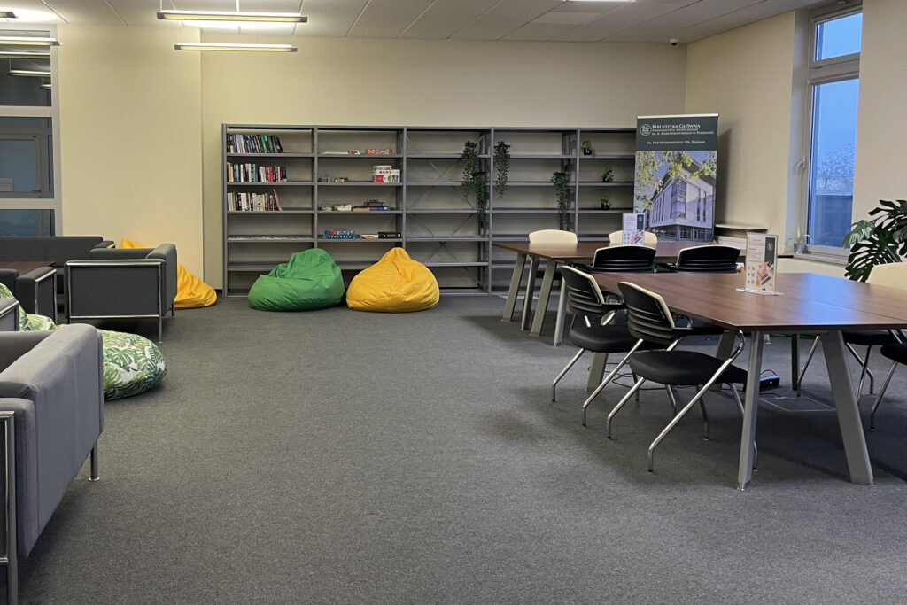 Duże pomieszczenie ze stolikami otoczonymi krzesłami i kanapami. Przy ścianie regał z książkami, kolorowymi pudełkami i roślinami. Na podłodze kilka dużych, kolorowych puf.