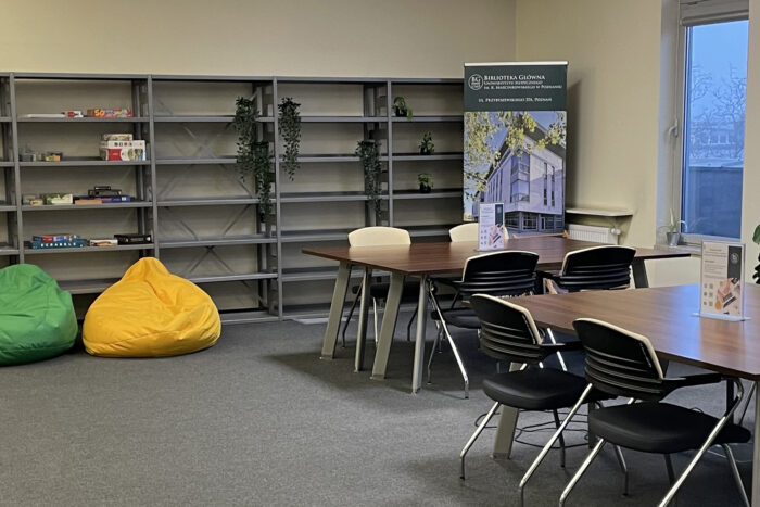 Duże pomieszczenie ze stolikami otoczonymi krzesłami. Pod ścianą regał z kolorowymi pudełkami i roślinami doniczkowymi. Przed nim dwie duże pufy: zielona i żółta