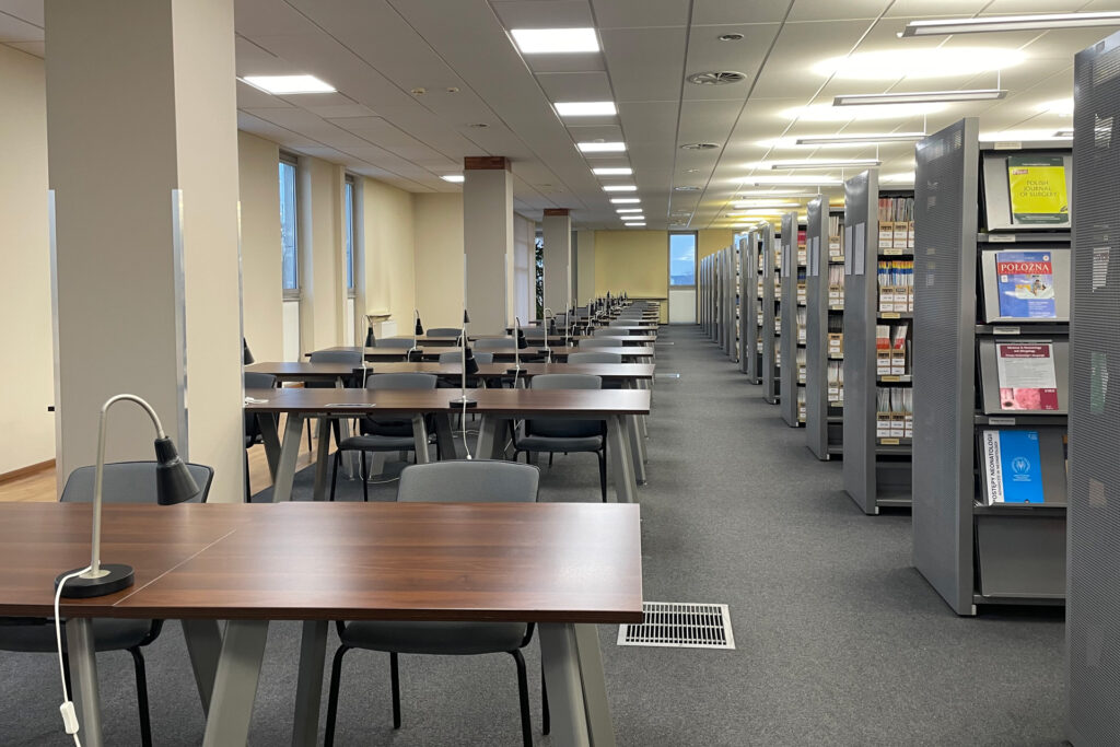Czytelnia Czasopism - rząd stolików z lampkami i krzesłami dla czytelników. Obok rząd regałów z czasopismami.