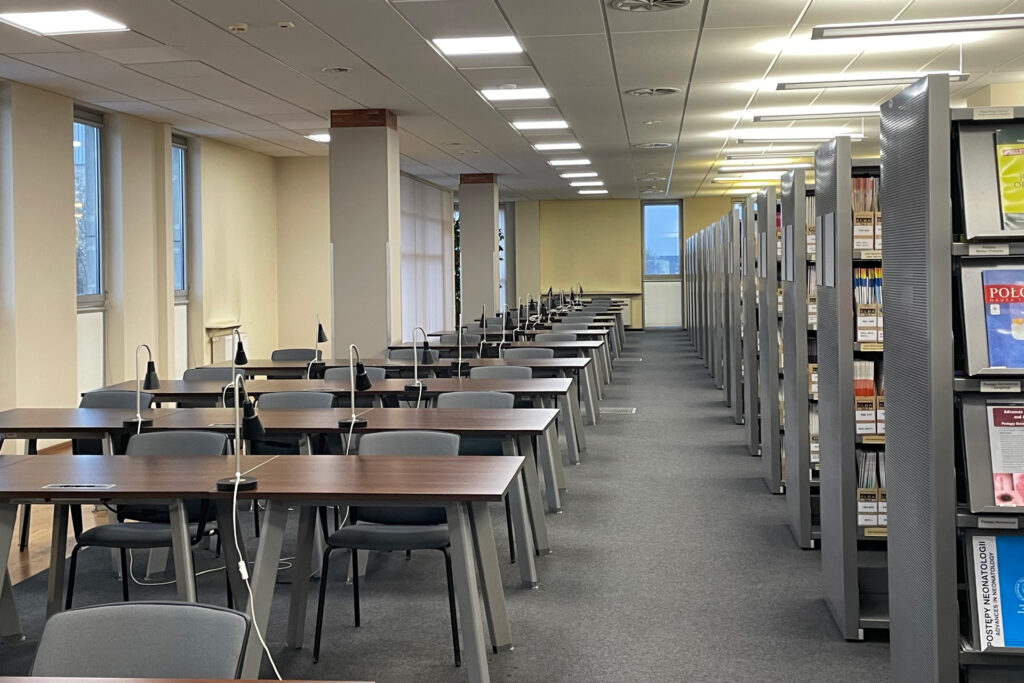 Czytelnia Czasopism - rząd stolików z lampkami i krzesłami dla czytelników. Obok rząd regałów z czasopismami.