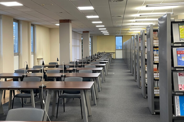 Czytelnia Czasopism - rząd stolików z lampkami i krzesłami dla czytelników. Obok rząd regałów z czasopismami.