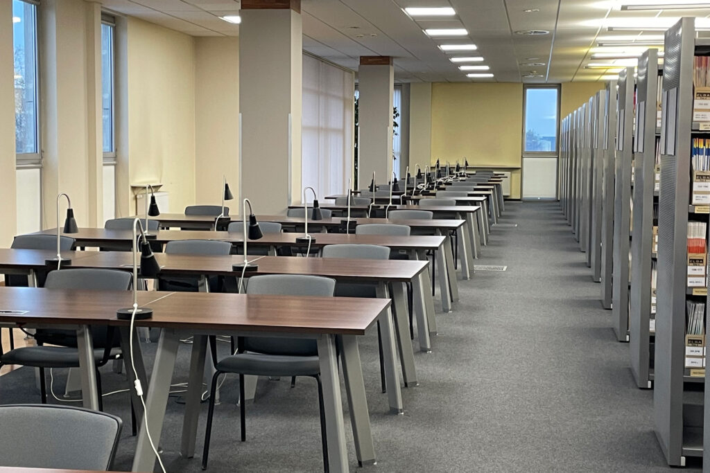 Czytelnia Czasopism - rząd stolików z lampkami i krzesłami dla czytelników. Obok rząd regałów z czasopismami.